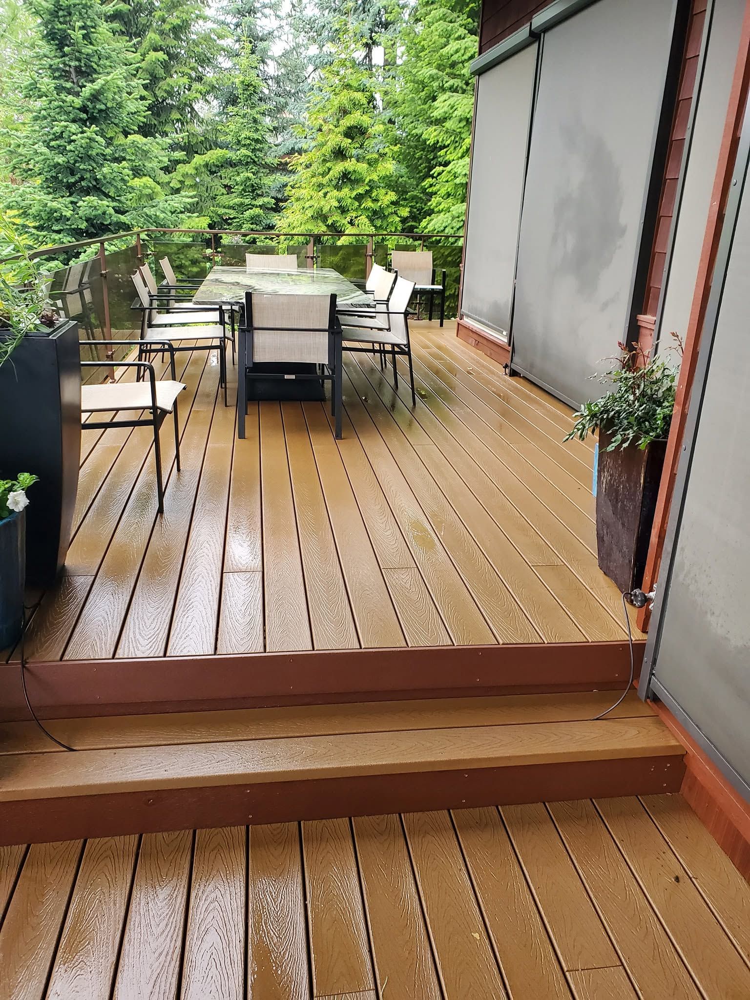 Wooden deck patio with outdoor dining table, chairs, and potted plants beside a house and trees.