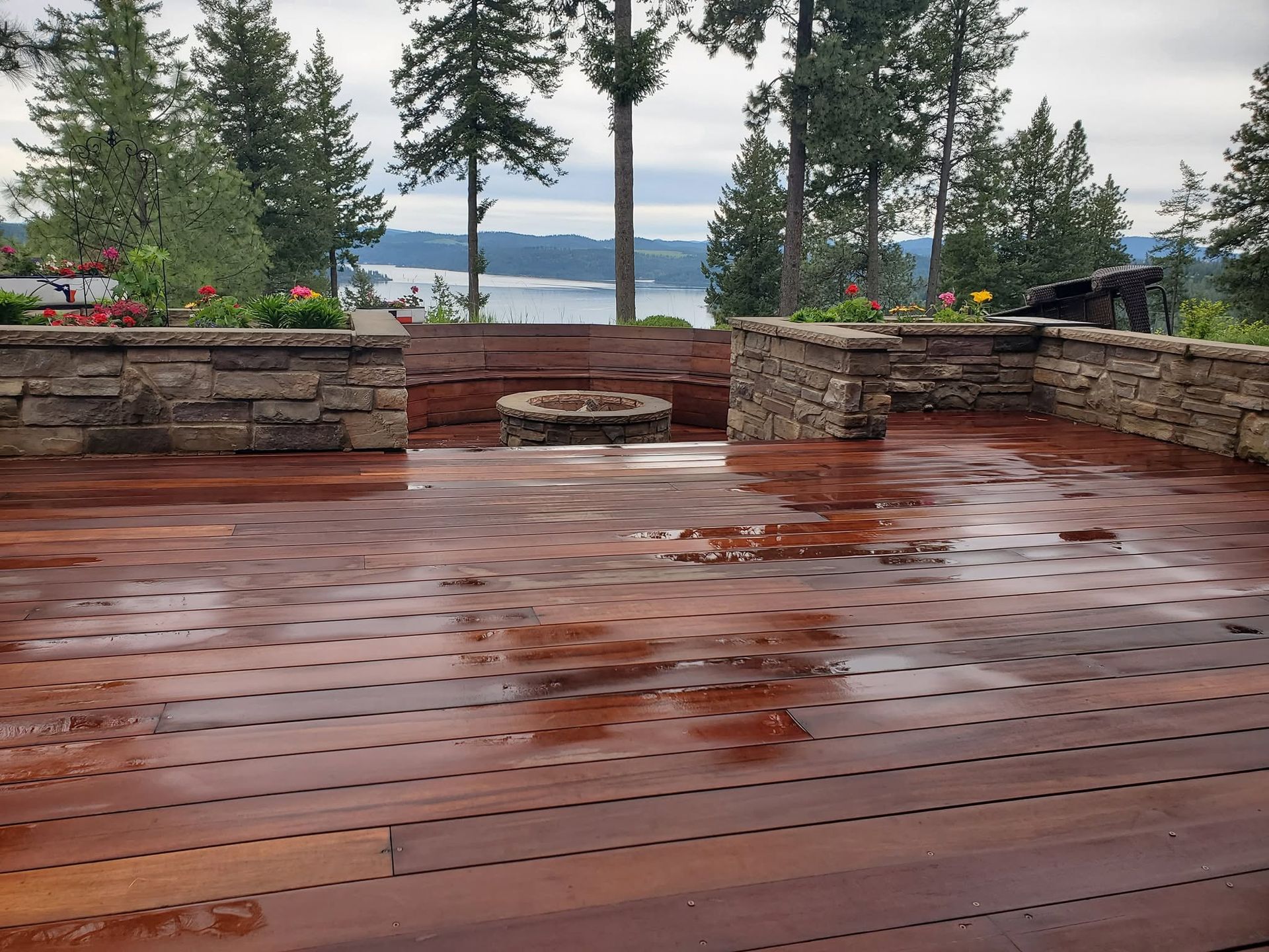 Rainy wooden deck with stone benches and tall trees overlooking a lake.