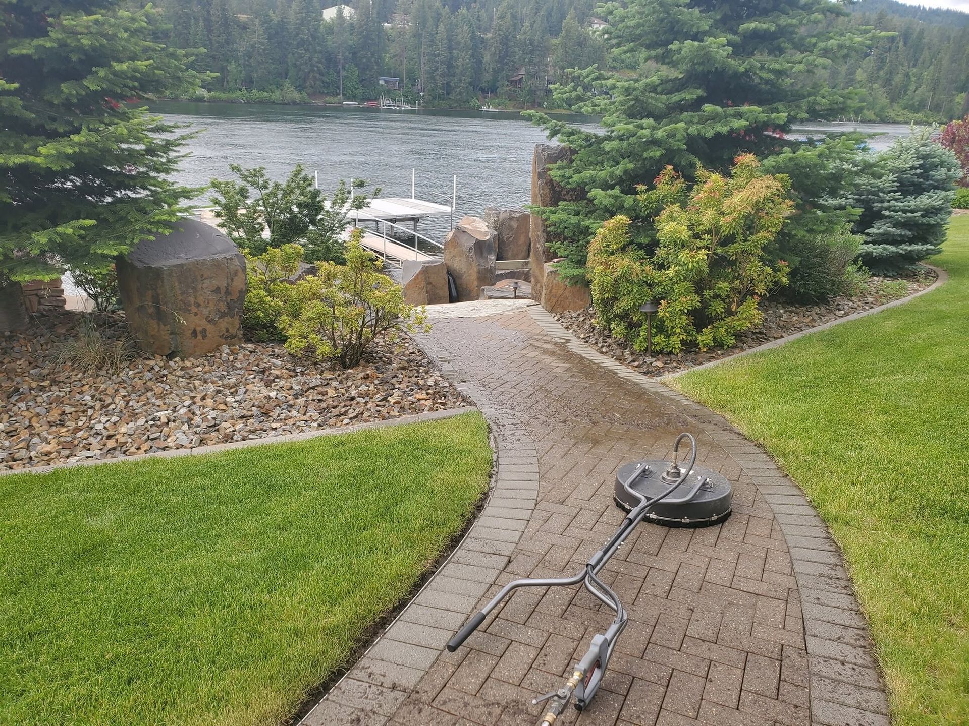 Curved brick path leading to a lakeside dock, with green lawn, rocks, and trees nearby