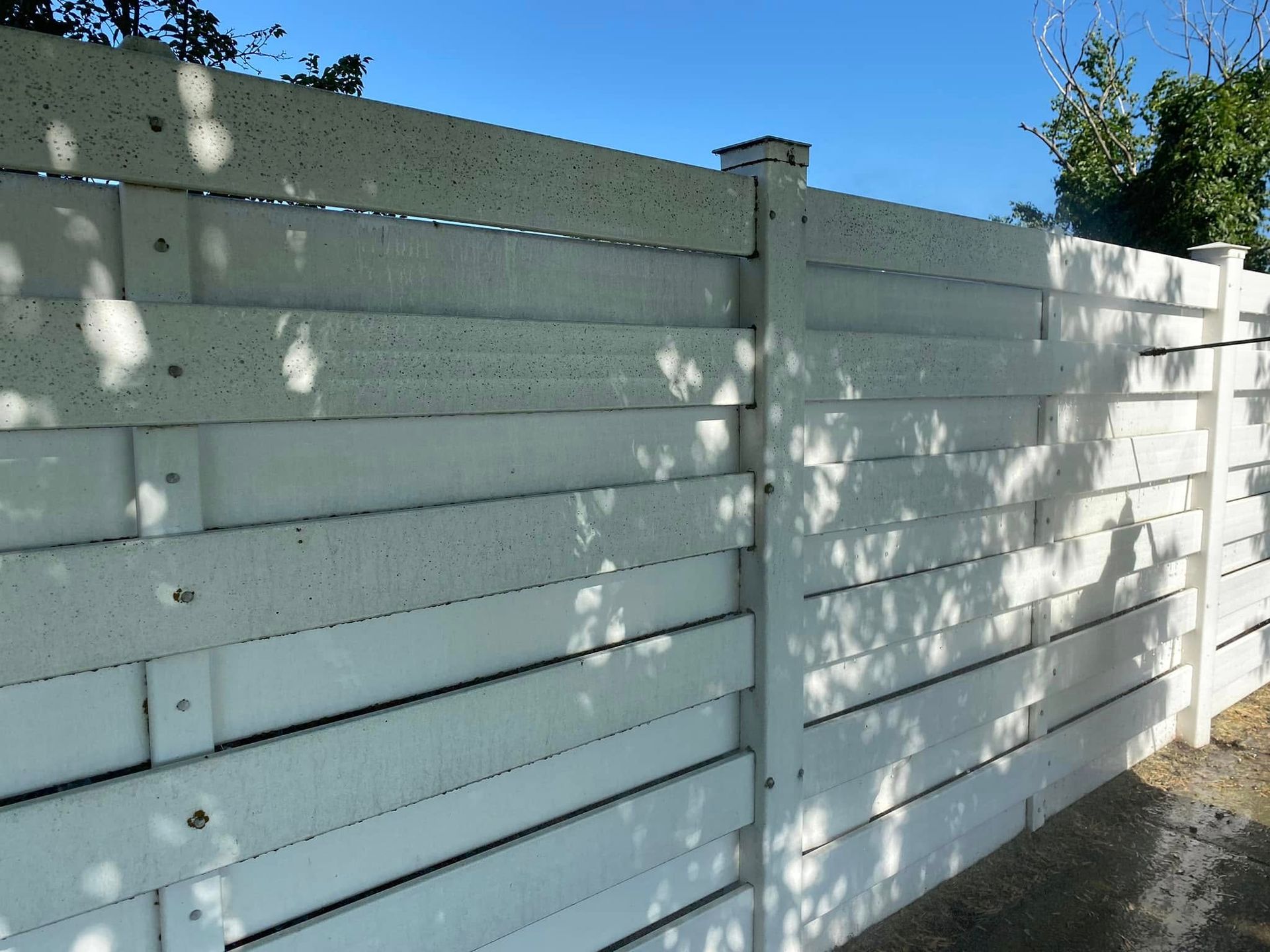 White wooden fence in dappled sunlight beside a paved path under a clear blue sky