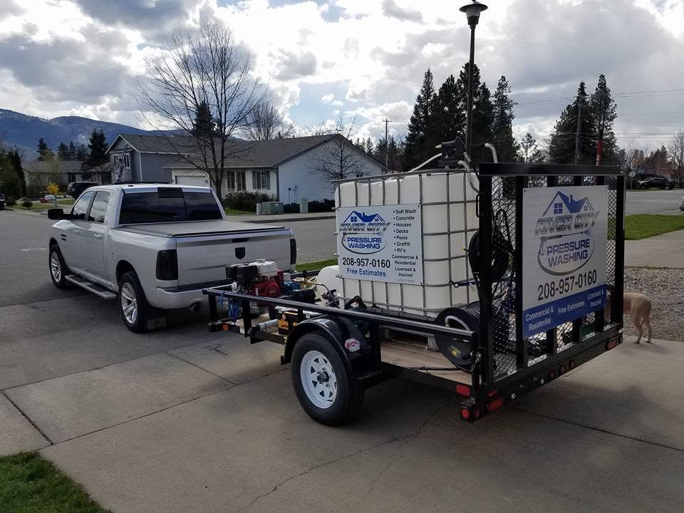 White pickup towing a small trailer carrying two large water tanks on a driveway, with houses and mountains in back.