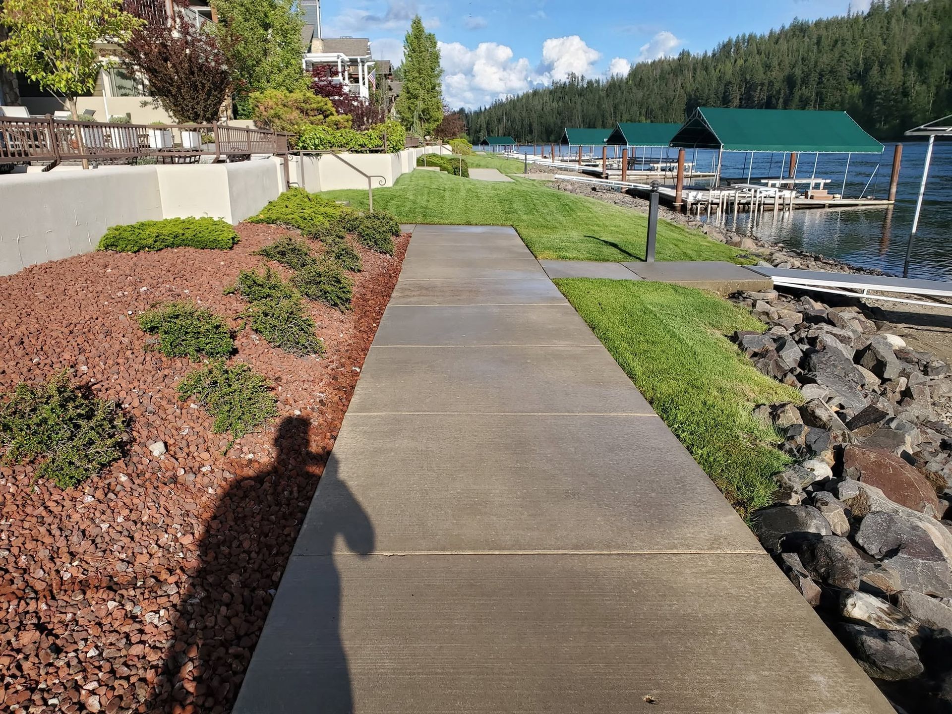 Concrete walkway beside marina, with red mulch, green landscaping, rocks, and blue-roofed docks under a sunny sky