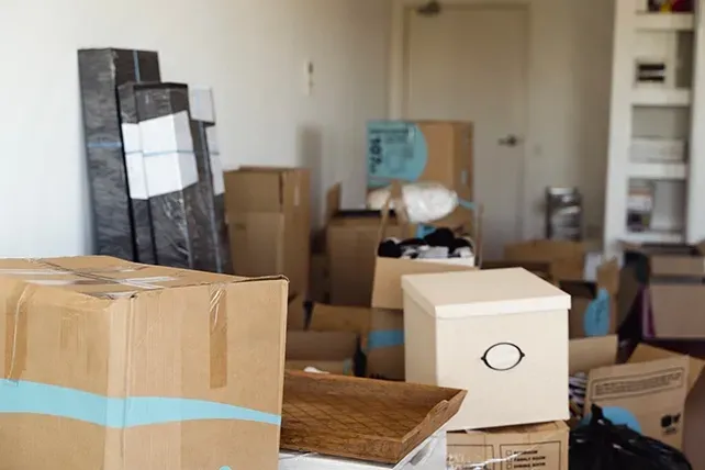 Stacked moving boxes in a room, with furniture and belongings scattered during a move.