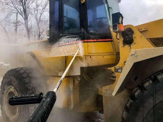 Worker pressure-washing a large yellow construction vehicle, spraying mud and dust off a tire outdoors