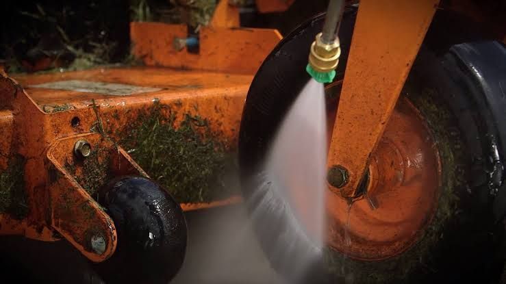 Orange lawn mower deck being sprayed with water, cleaning grass clippings from the wheel and underside.