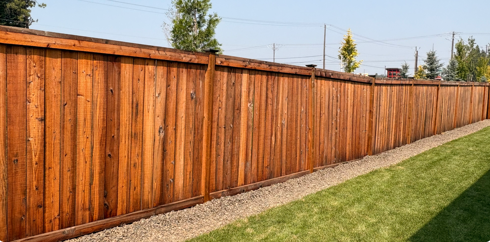 Long wooden privacy fence along a gravel edge beside a green lawn under a clear sky
