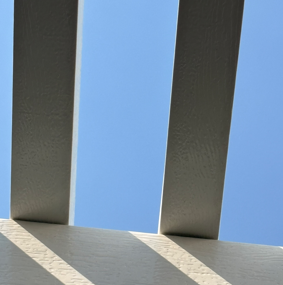 Blue sky framed by dark beams over a white wall with diagonal shadows