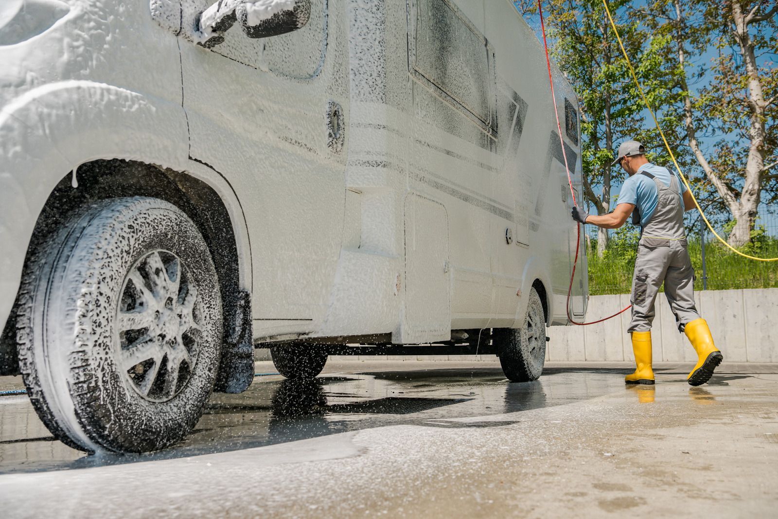 Person pressure-washing a white vehicle outdoors with water spraying and yellow boots visible