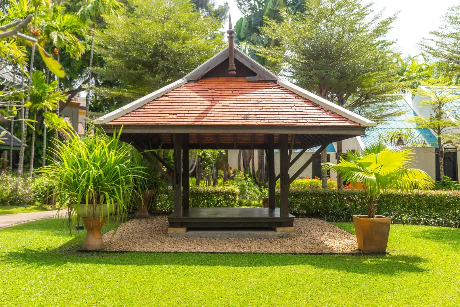 Small open-sided gazebo with a red roof in a lush green garden, surrounded by plants and trees.