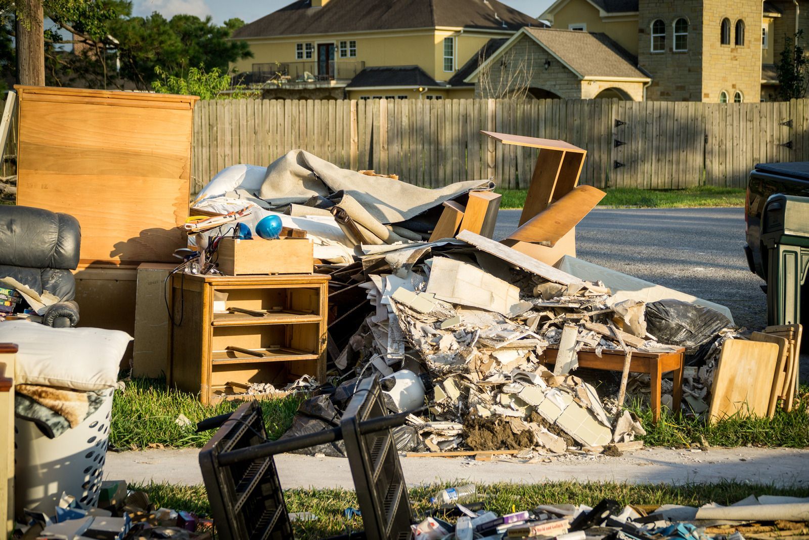 Trash and debris piled in a yard beside wooden fencing, with a dresser and broken materials scattered around.