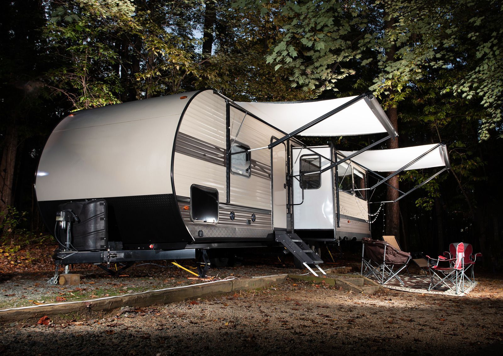 Camouflaged travel trailer with awning and camp chairs in a wooded campsite at night