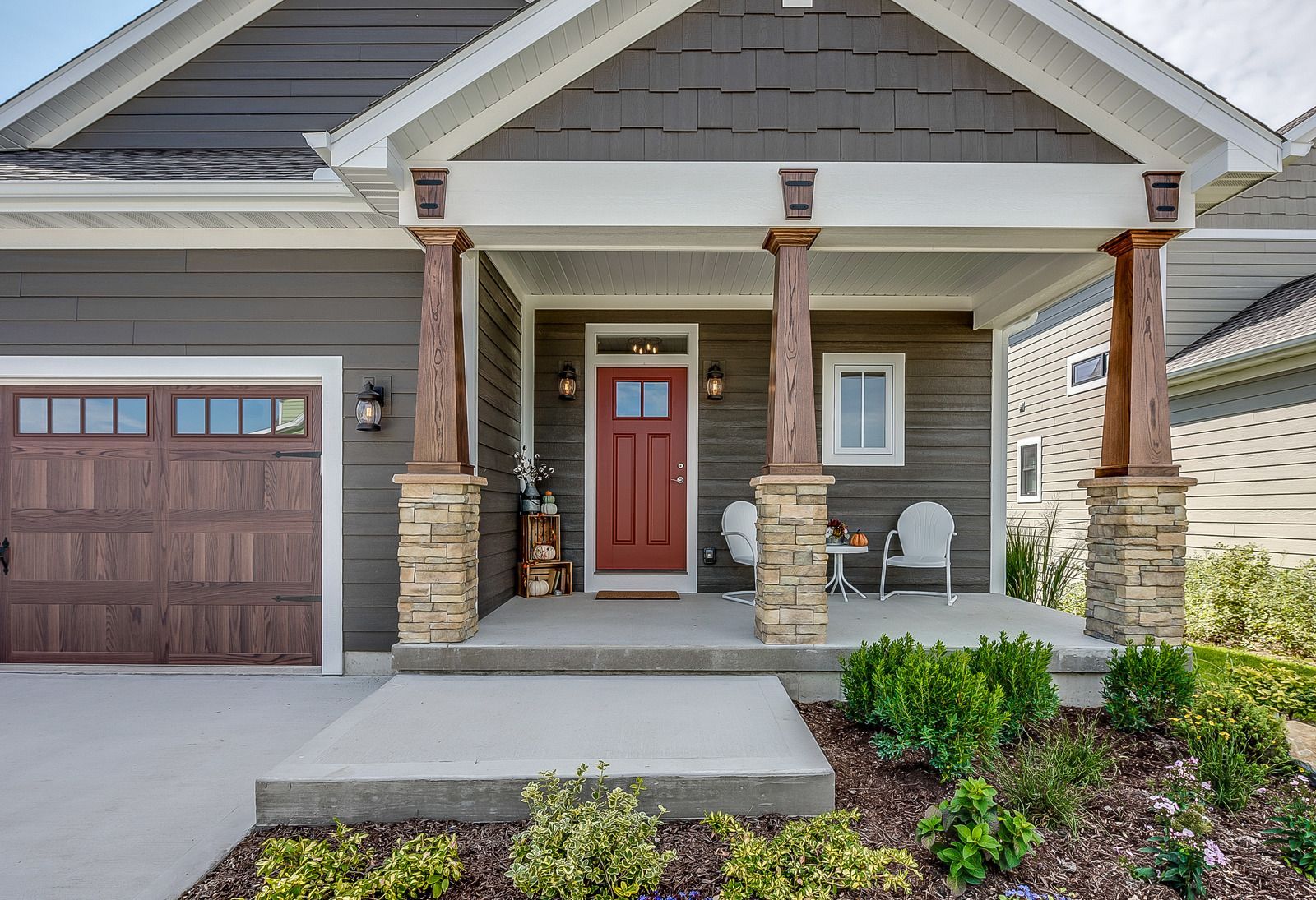 Front porch of a gray house with red door, stone columns, garage, and small landscaped garden.