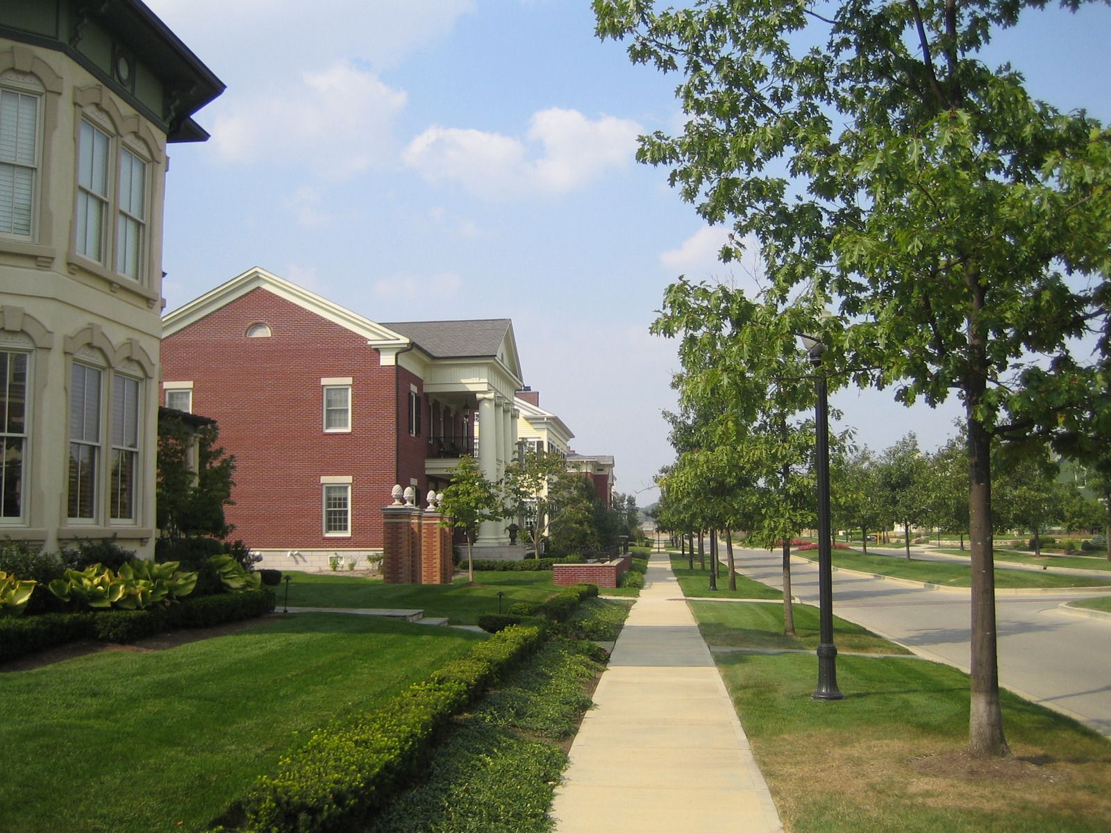 Residential street with apartment buildings, sidewalk, trees, and a quiet road under a partly cloudy sky