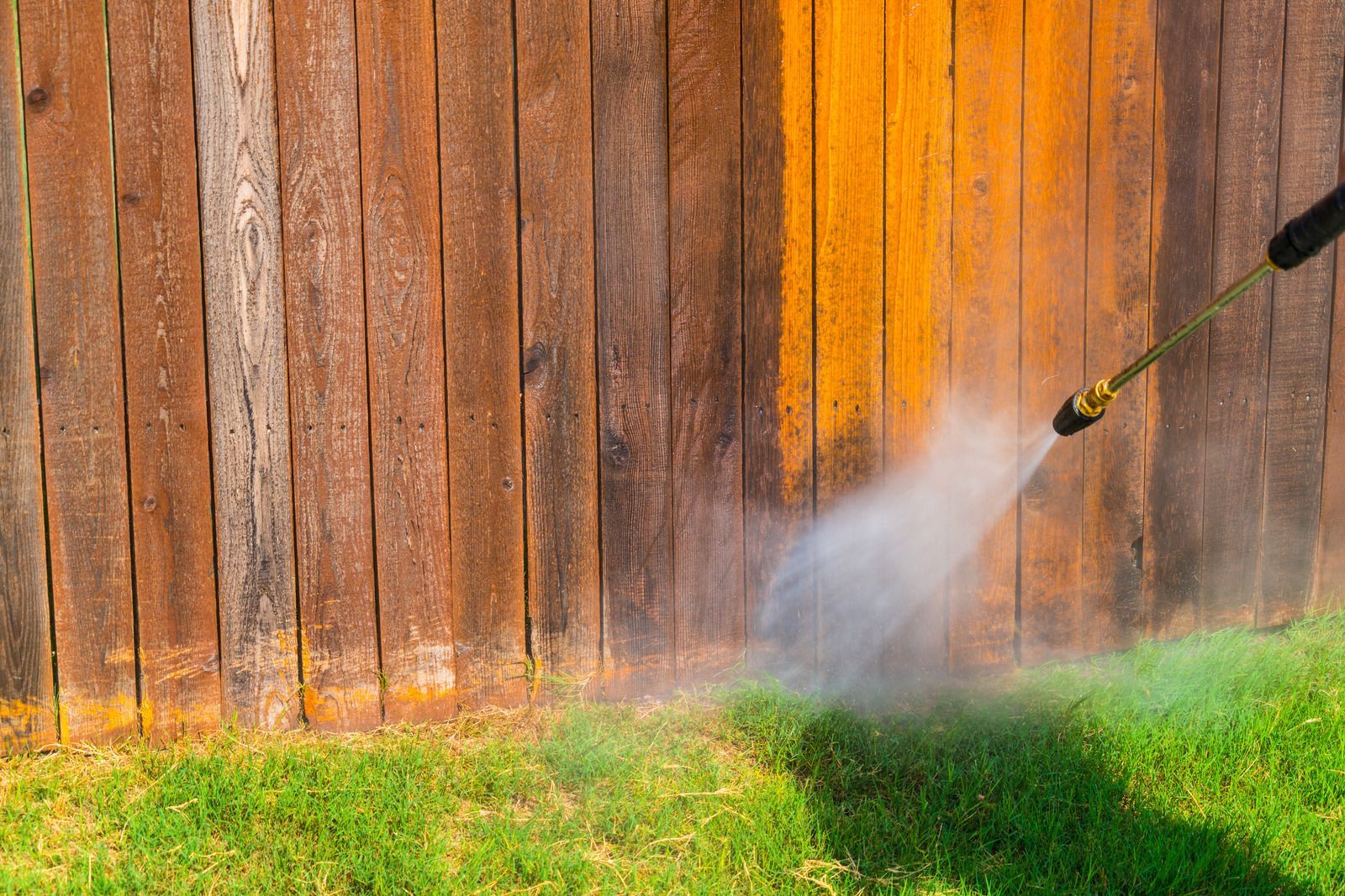 Pressure washing a wooden fence, with water spray over green grass.
