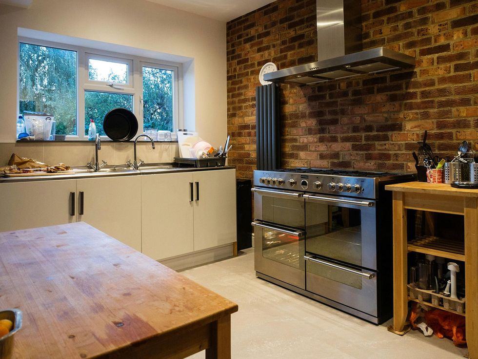 A kitchen with a large stainless steel range cooker, a brick feature wall, light cabinets, and a wooden table in the front.
