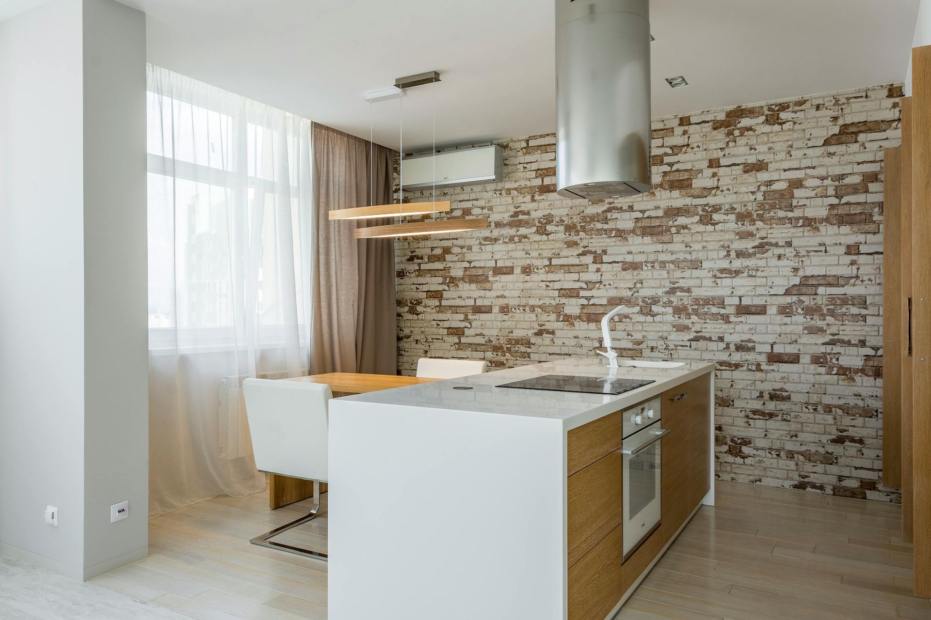 A modern kitchen featuring a white island with a built-in oven, fronted by a brick-patterned wall and a dining area.