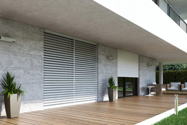Esterno di un edificio moderno con persiane bianche, una terrazza in legno e piante in vaso.