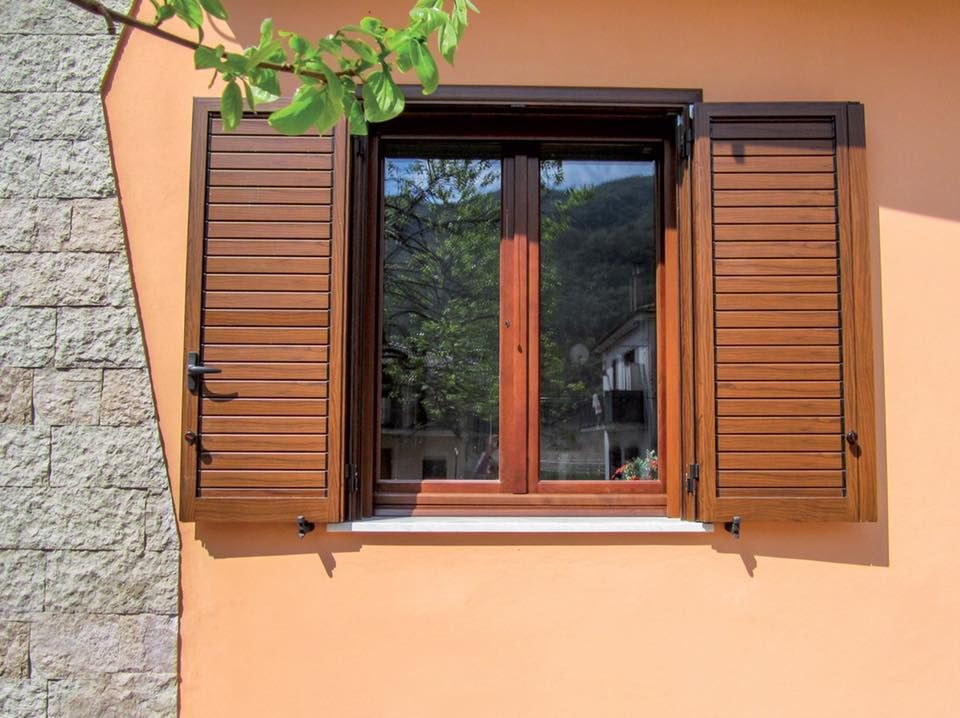 Finestra con persiane in legno marrone che si apre su una parete color pesca, che riflette il verde.