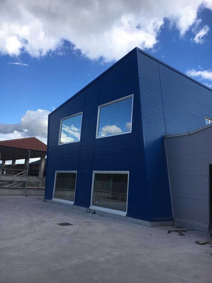 Edificio industriale blu e grigio con finestre riflettenti contro un cielo azzurro nuvoloso.