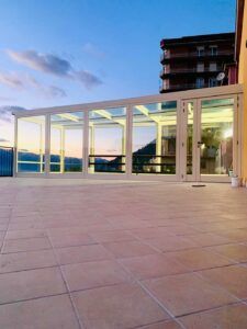 Terrazza con pavimento piastrellato, struttura con pareti in vetro e edificio sullo sfondo al tramonto.