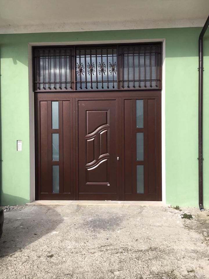 Porta in metallo marrone con motivo decorativo, pannelli in vetro e finestra in ferro battuto nella parte superiore di un edificio verde.