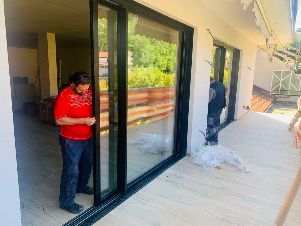 Un uomo con una camicia rossa sistema una porta scorrevole in vetro su un balcone. Un'altra persona sullo sfondo.