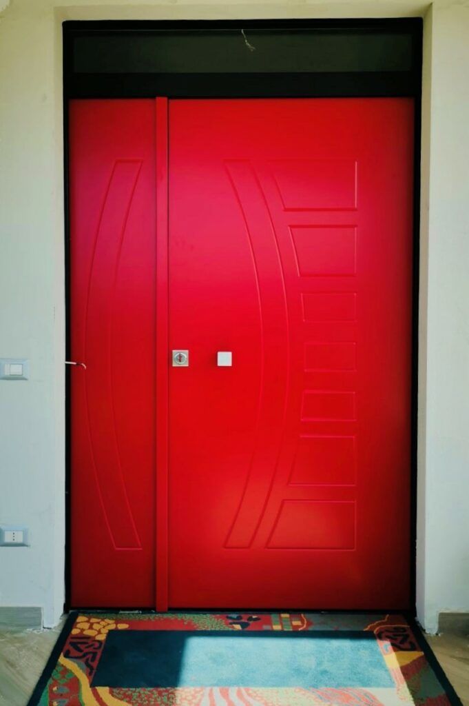 Porta doppia rosso brillante con disegno geometrico, inserita in una cornice nera; zerbino in primo piano.