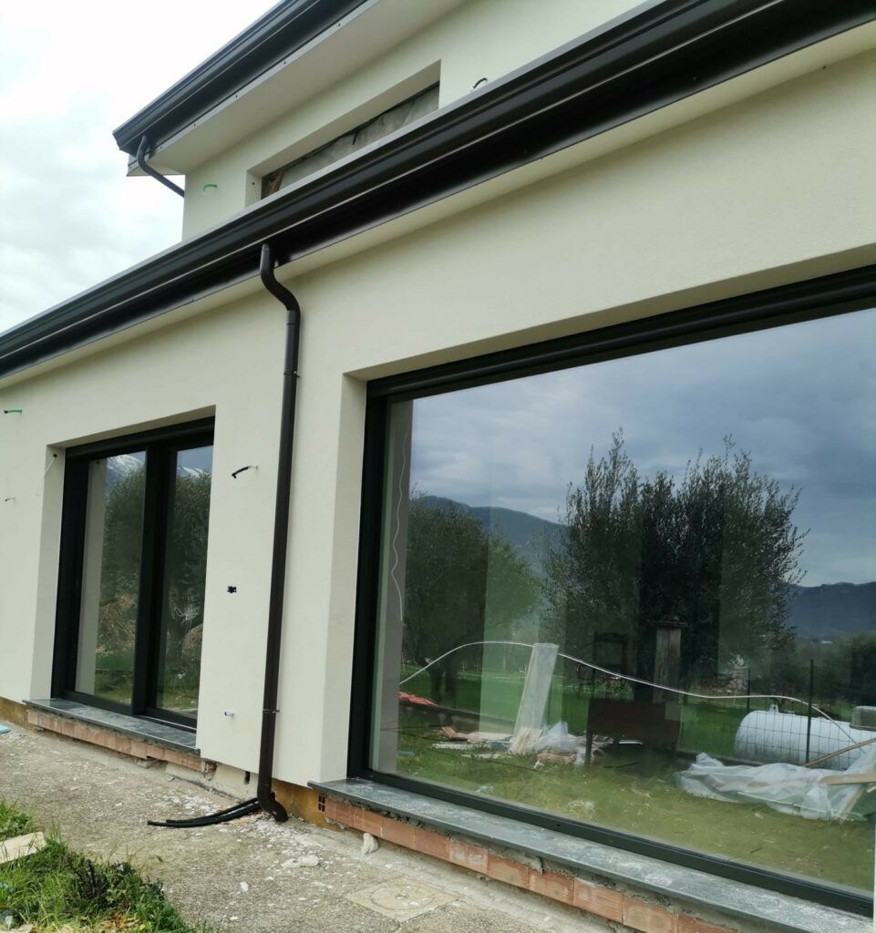Vista esterna di una moderna casa bianca con grandi finestre, infissi neri e grondaie, che riflette il verde e un cielo nuvoloso.