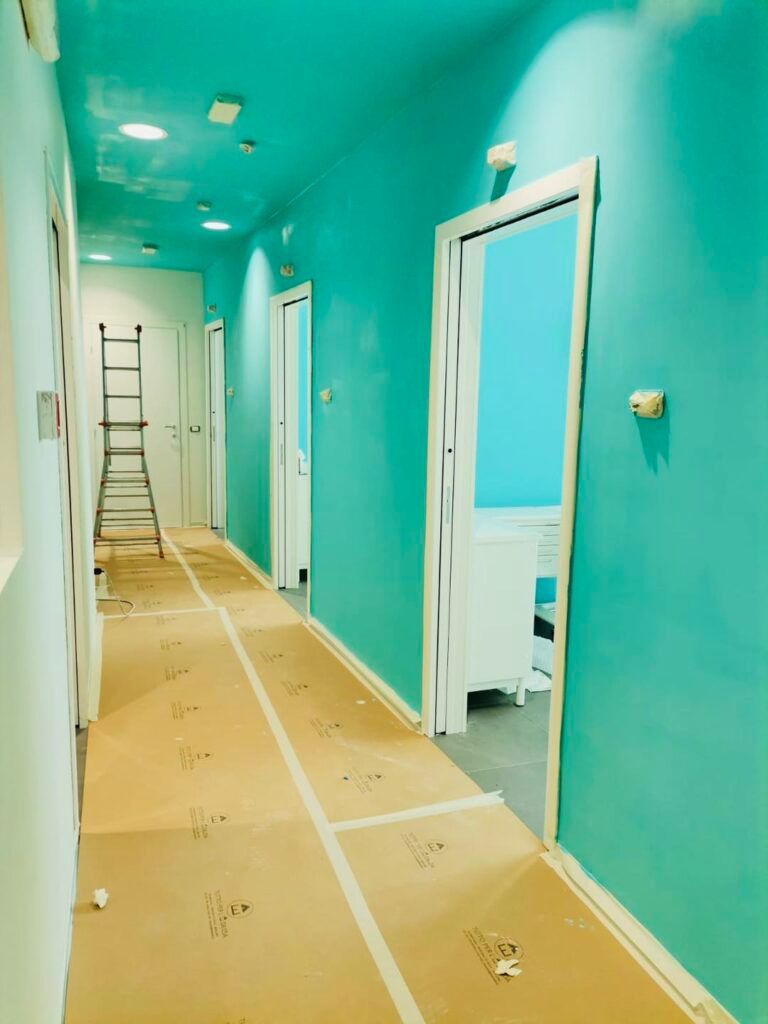 Lungo corridoio dipinto di turchese. Porte e soffitto sono bianchi. Una scala è appoggiata verso la fine.