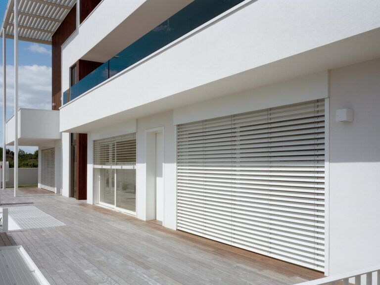 Edificio moderno bianco con persiane a stecche, terrazza in legno e balcone in vetro.