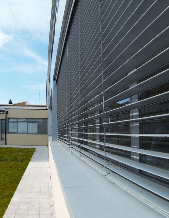 Esterno di un edificio moderno con persiane metalliche orizzontali che ombreggiano le finestre. Erba verde e cielo azzurro.