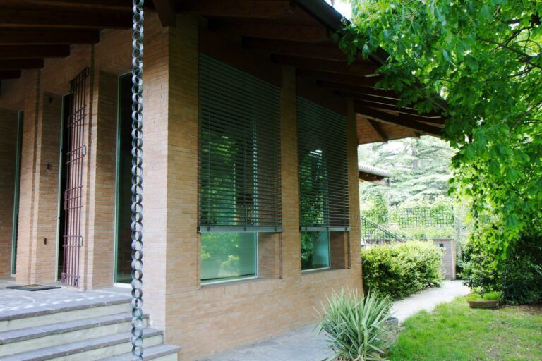 Casa color marrone chiaro con finestre coperte da persiane; gradini che conducono all'ingresso; la struttura è circondata dal verde.