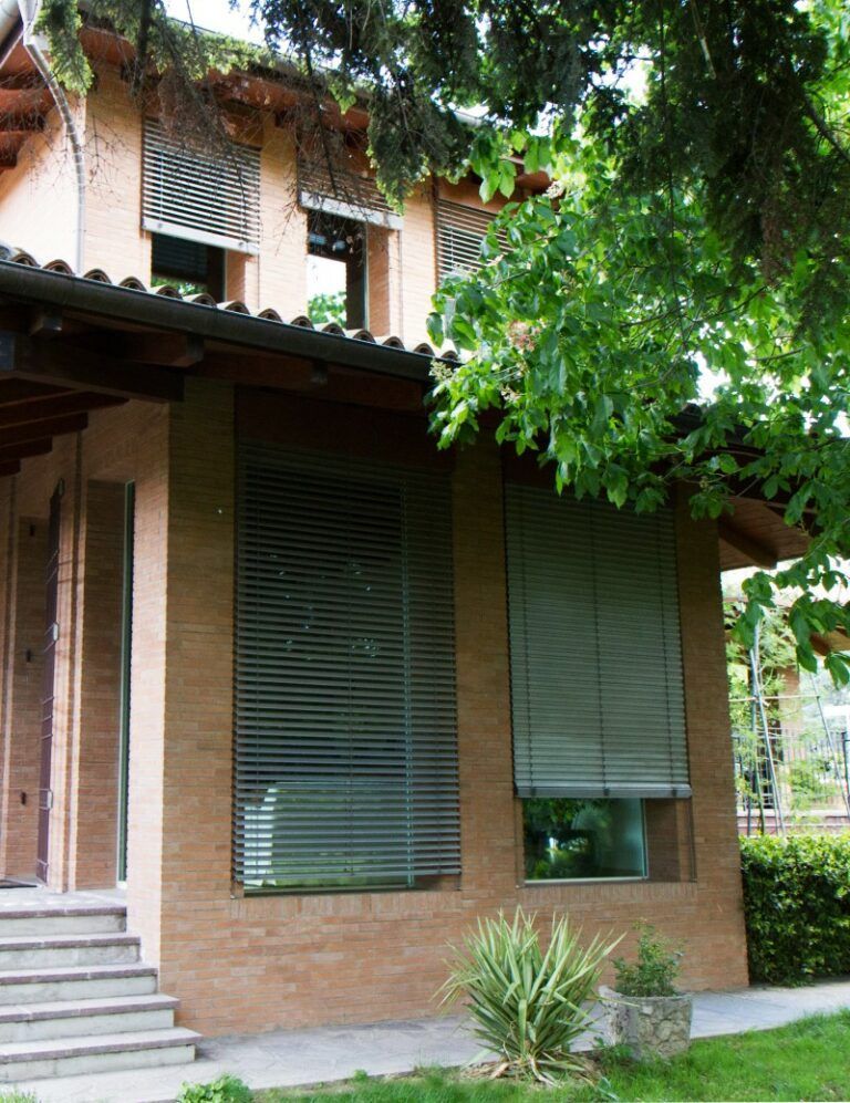 Esterno dell'edificio con facciata in mattoni, finestre con persiane e alberi sporgenti.