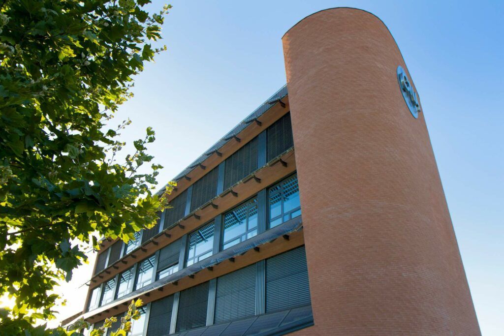 Edificio in mattoni con torre cilindrica e finestre orizzontali contro un cielo azzurro, rami di alberi in primo piano.