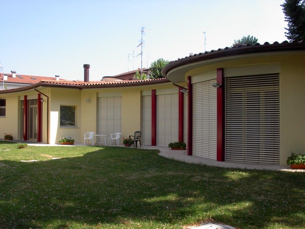 Edificio giallo con dettagli rossi, facciata curva, persiane e prato verde.