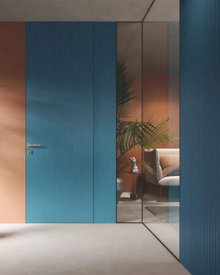 La porta blu e la parete in vetro rivelano un interno con una sedia e una pianta, su pareti color arancio e pesca.