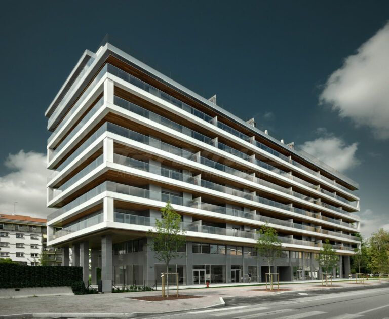 Edificio moderno a più piani con balconi in cemento bianco e vetro, pilastri di sostegno grigi e cielo azzurro.