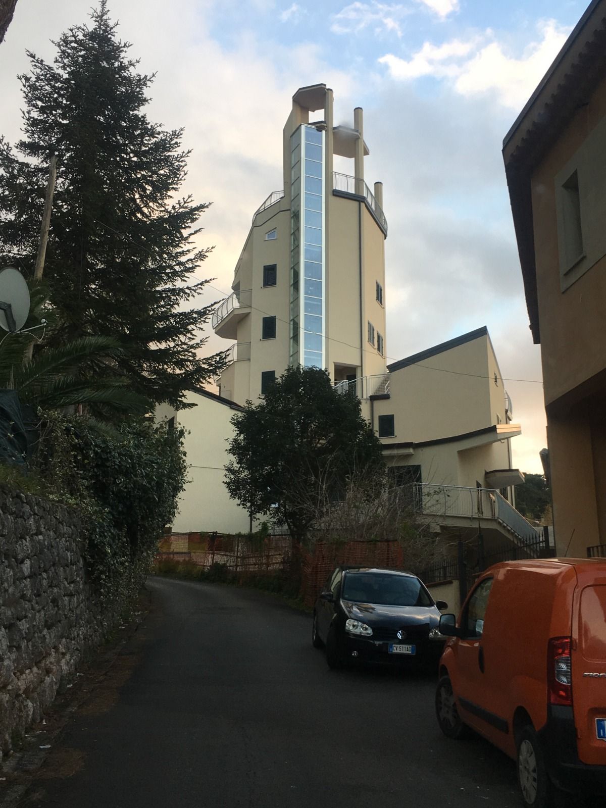 Edificio alto con ascensore esterno in una strada stretta; auto parcheggiate in primo piano, alberi su entrambi i lati.