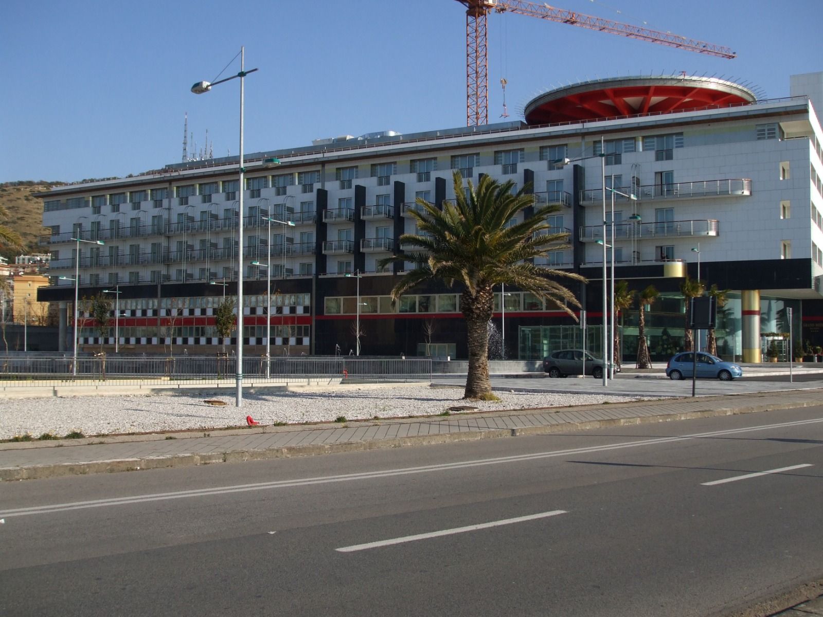 Edificio moderno con accenti rossi, una palma e una gru; ambiente soleggiato all'aperto.