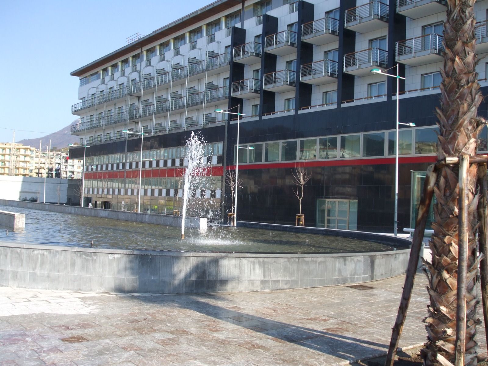 Fontana in una vasca circolare di fronte a un edificio moderno con balconi; palma in primo piano.