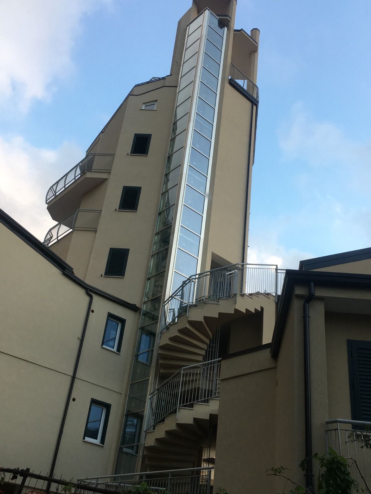 Esterno di un edificio con ascensore in vetro e scala a chiocciola. L'edificio è beige, con finestre e balconi scuri.