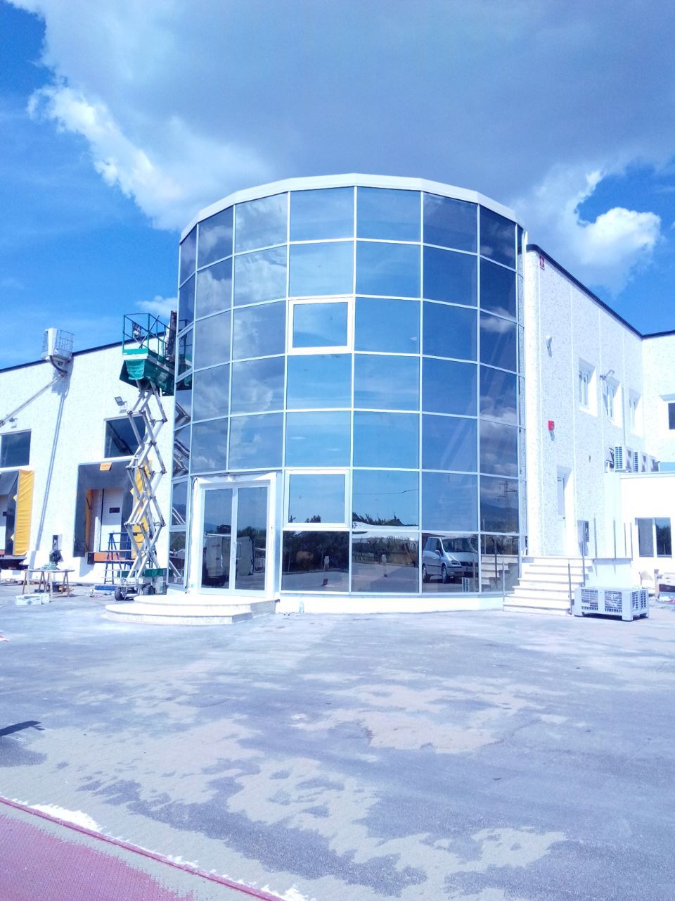 Edificio moderno con facciata curva in vetro che riflette il cielo azzurro e le nuvole. Sono presenti attrezzature edili.