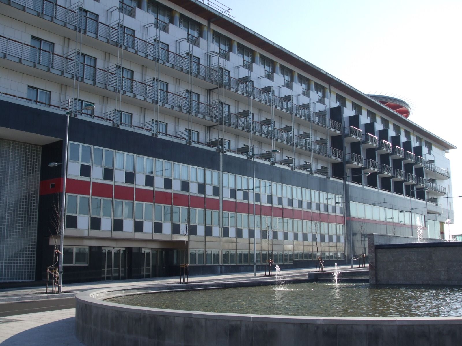 Moderno edificio a più piani con balconi, facciata nera, rossa e bianca e una fontana.