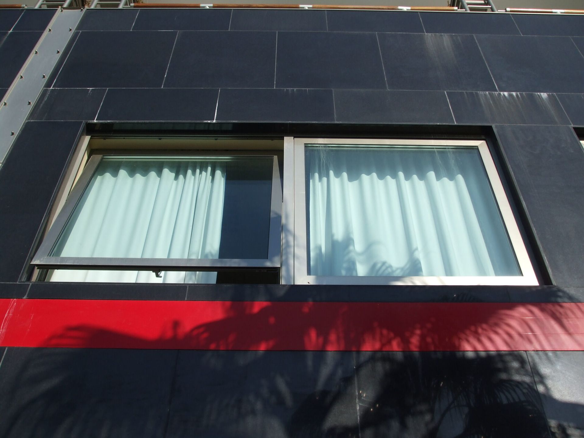 Due finestre su un edificio nero con una striscia rossa. Una finestra è aperta, entrambe con tende azzurre.