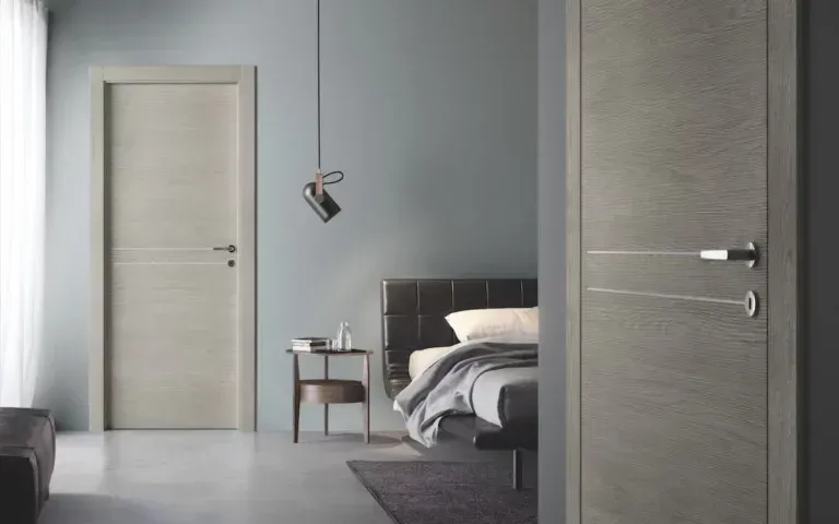 Interno della camera da letto con due porte in legno grigio chiaro, letto e comodino contro una parete blu tenue.