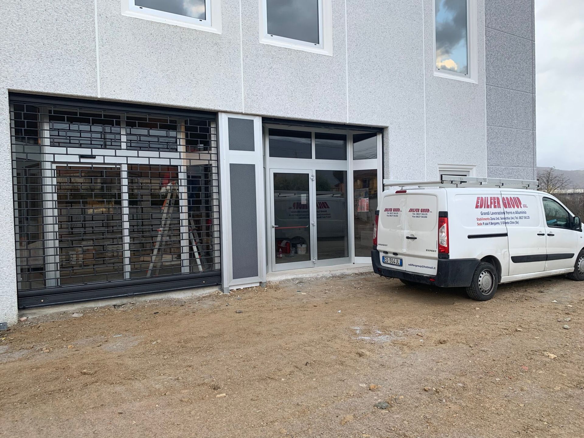 Esterno di un edificio con un furgone bianco parcheggiato all'esterno. L'edificio ha un ingresso con porta a vetri e un cancello di sicurezza.