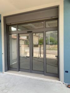 Porte scorrevoli automatiche in vetro con telaio grigio, montate sulla facciata di un edificio, che riflettono l'area circostante.