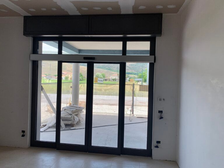 Porte scorrevoli in vetro con apertura automatica, telaio grigio, che conducono a un'area esterna.