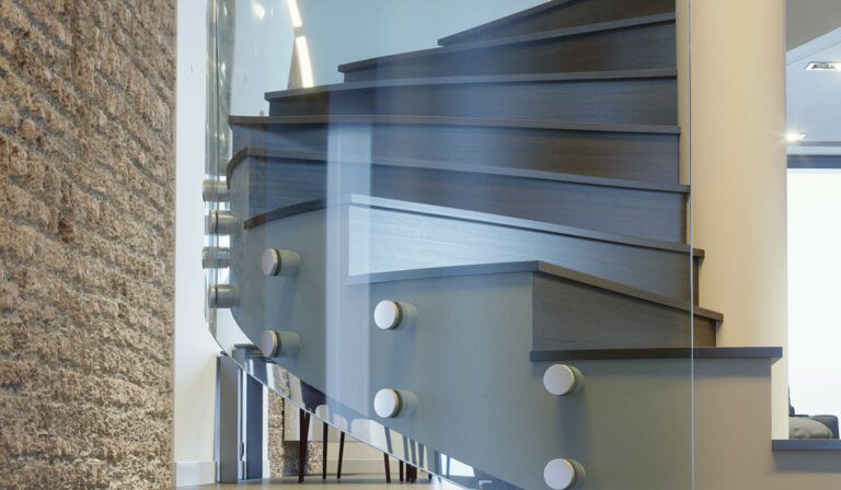 Scala moderna con pannello in vetro e gradini grigi. Muro di mattoni a sinistra, pilastro bianco a destra.