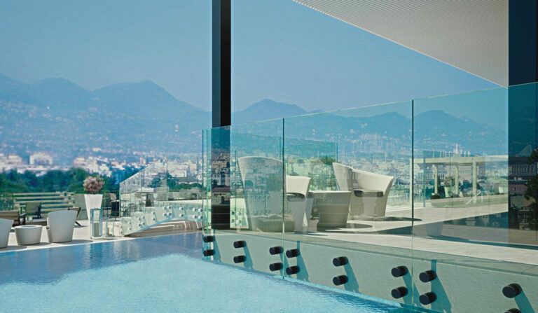Piscina sul tetto con vista sulla città e sulle montagne, ringhiera in vetro, mobili bianchi. Giornata luminosa e soleggiata.